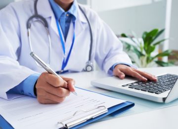 hands-unrecognizable-female-doctor-writing-form-typing-laptop-keyboard