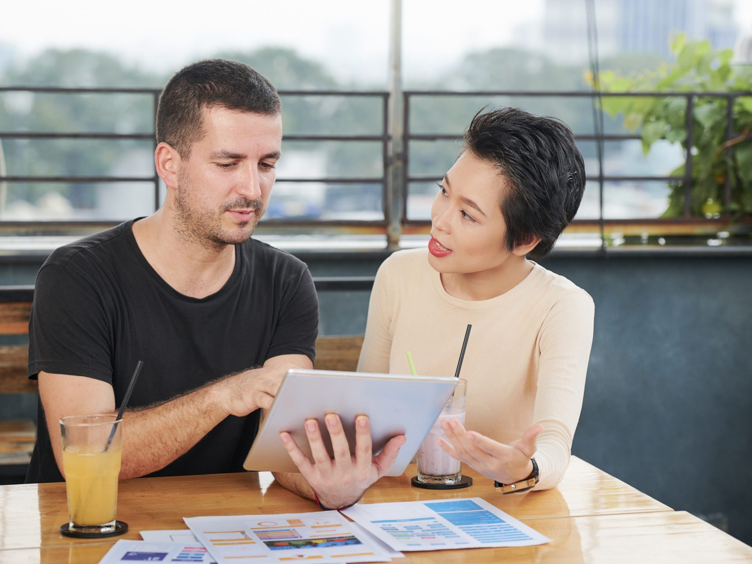 Young businessman using digital tablet and working online while his Asian colleague talking to him during their meeting in cafe