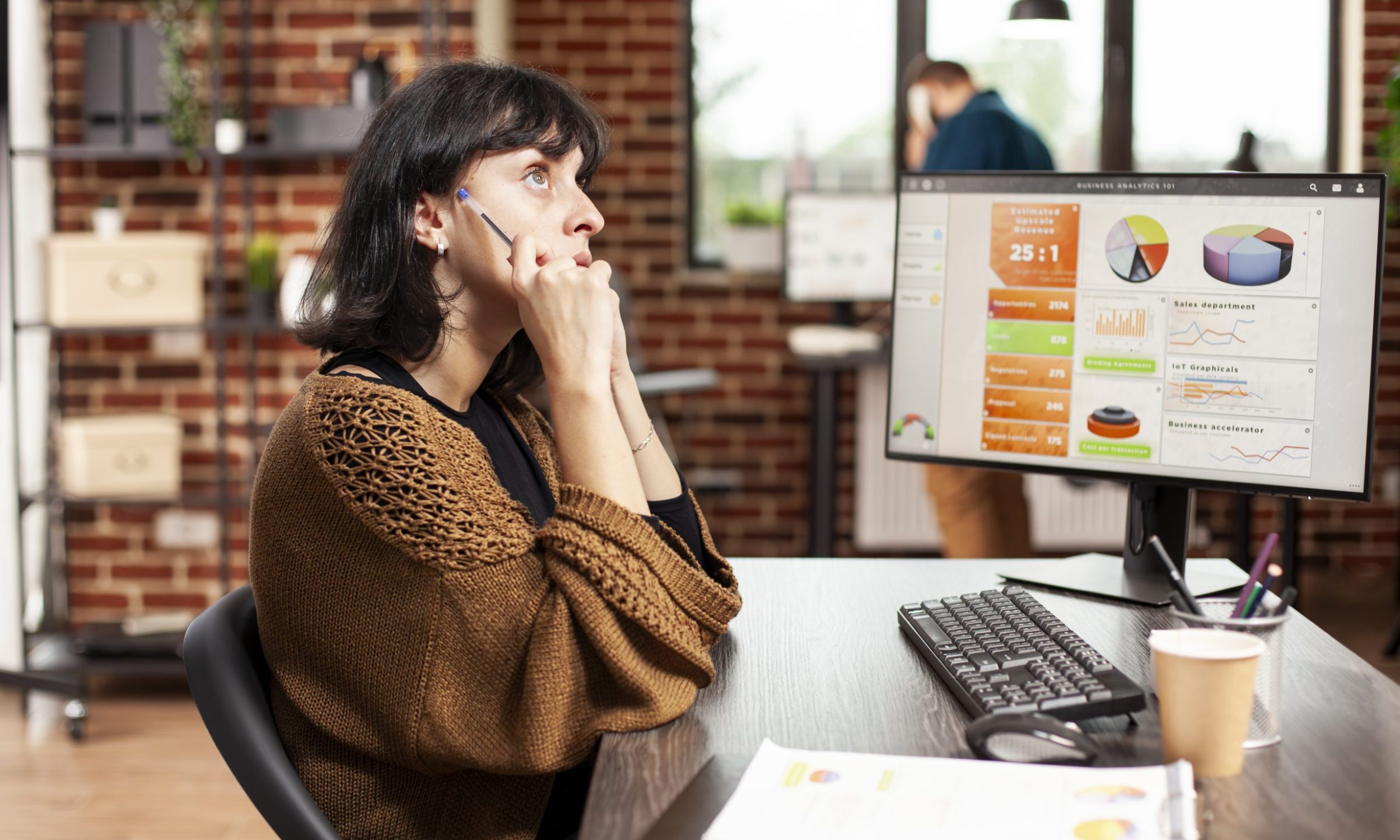 Female employee sits at office desk having brainstorming session as desktop monitor displays market data. Woman with pen, deeply thinking about project ideas for startup company at brick wall room.